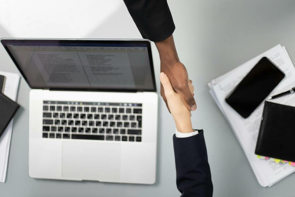 Zwei Personen geben sich über Schreibtisch mit Laptop und Unterlagen die Hand, Symbolfoto für Spezialservices für Steuerberater.